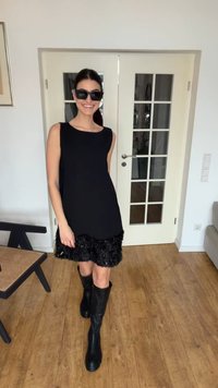 Kvinne som smiler, iført svart ermeløs kjole med teksturert kant, svarte knelange støvler og solbriller, stående innendørs ved hvite dobbeltdører.