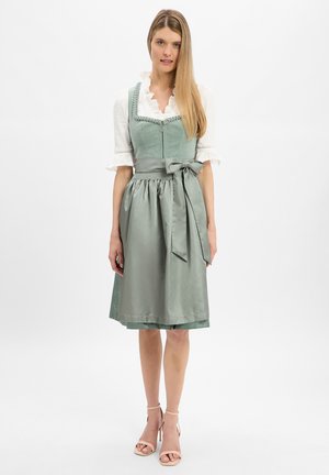 Woman wearing a light green traditional dress with apron and white blouse, standing on a white background, beige high-heeled sandals.