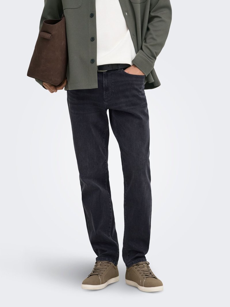 Personne portant un jean foncé, une veste vert olive, une chemise blanche, des baskets marron, et tenant un grand sac fourre-tout marron dans une main.