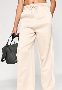 Bež sproščene trenirke hlače z elastiko v pasu, vrvico za zategovanje in belimi stranskimi črtami. V roki drži temno torbo.