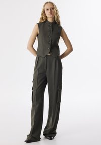 Gilet sans manches vert avec des boutons sur le devant et un col, associé à un pantalon cargo à jambes larges comportant des poches latérales et un tissu texturé.