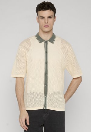 Mann trägt ein beigefarbenes, kurzärmeliges Strickhemd mit Knopfleiste und olivgrünem Besatz, kombiniert mit schwarzen Hosen, steht vor hellem Hintergrund.