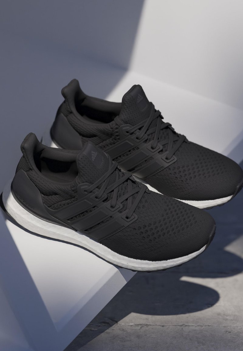 Sapatos de desporto pretos com um cabedal de malha texturizada, apresentando três riscas pretas e uma sola intermédia branca. O design inclui um ajuste justo e um fecho com atacadores.