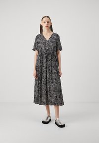 Musta mekko valkoisilla pistekuvioilla, lyhyet hihat, V-kaula-aukko, vyötäröä korostava, nilkkapituinen. Mallilla on päällään hopeiset platform-kengät.