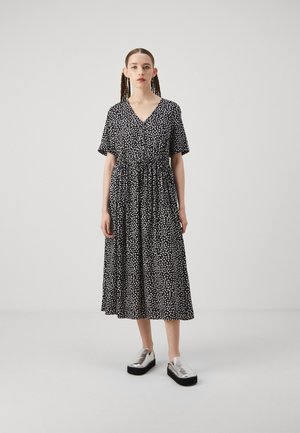 Black dress with white dotted pattern, short sleeves, V-neck, cinched waist, ankle-length. Model wearing silver platform shoes.