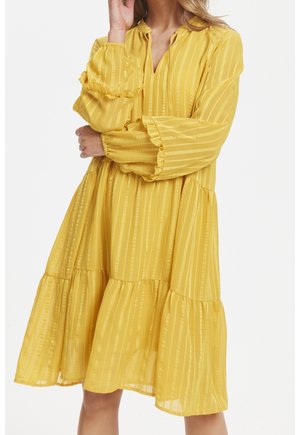 Žluté vrstvené šaty z lehké tkaniny s vertikálními pruhy. Mají dlouhé rukávy s volánkovými manžetami a jemný výstřih do V.