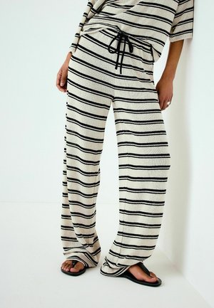 Kvinde iført løse cremefarvede strikkede bukser med sorte vandrette striber og snøre, kombineret med sorte klipklappere, stående op ad en hvid væg.