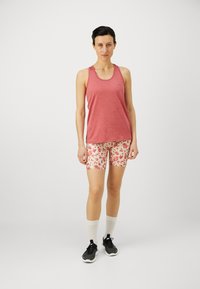 Canotta senza maniche rossa mélange, abbinata a pantaloni corti crema con stampa floreale. La modella indossa sneakers nere e calze bianche. Design semplice.