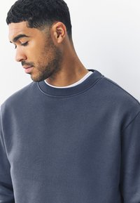 Tengerészkék pulóver kerek nyakkivágással és bordázott mandzsettákkal; puha, sima tapintású anyagból készült. Alatta fehér ing rétegezve.