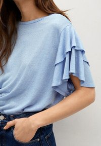 Lyseblå kortærmet bluse med lagdelt flæse-design ved ærmerne. Blød, tekstureret stof med en afslappet pasform, kombineret med mørk denim.