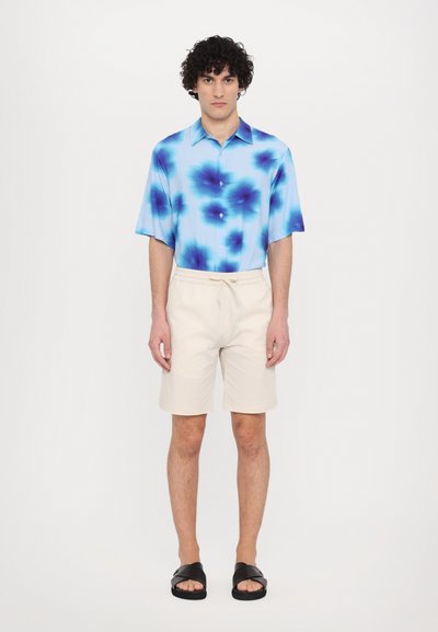 Cămașă cu mânecă scurtă de culoare albastru deschis, cu model tie-dye în nuanțe de albastru închis, asortată cu șorturi bej cu șnur și sandale negre tip slide.