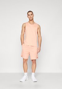 Sæt bestående af en ferskenfarvet tanktop og shorts lavet af blødt, åndbart stof; har rund halsudskæring, sideslids og logo-detalje. Bæres med hvide sportssko.