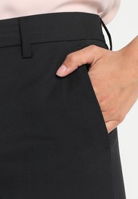 Černé kalhoty na míru s hladkým materiálem, rovným střihem a bočním kapsou. Ruka spočívá v kapse, což ukazuje na čistý design.
