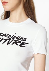 Vit bomulls t-shirt med rund hals, som har djärv svart typografi som säger "versace jeans FUTURE" och krusade ärmslut.