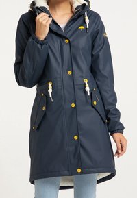 Sateen navy veske med hette, hvit fuskepelsfôr, gule knapper og snorer. Har to sidelommer og justerbare mansjetter.