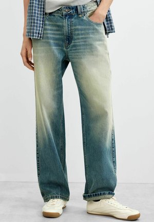 Person trägt ausgewaschene blaue Jeans mit weitem Bein und hat die Hand in der Tasche, weiße Sneakers, graues T-Shirt und offen getragenes blau kariertes Hemd, steht auf grauem Boden.