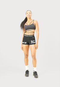 Donna atletica in piedi, con indosso un reggiseno sportivo nero con bretelle, pantaloni corti Nike Pro neri, calze bianche Nike e sneakers nere su uno sfondo chiaro e semplice.