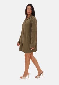 Olivengrøn skjortekjole med knapper lavet af tekstureret stof, med lange ærmer, løs pasform og sideslids. Tilbehør: beige hælede sandaler.
