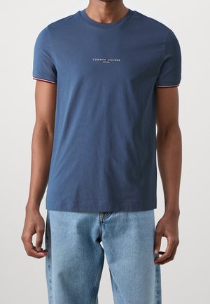 Mand iført en blå Tommy Hilfiger T-shirt med lille logo tekst og lyseblå jeans, stående mod en ensfarvet hvid baggrund.