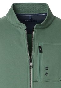 Veste zippée verte avec col montant, tissu texturé, poche zippée latérale et deux boutons pression. Étiquette : Casa Moda.