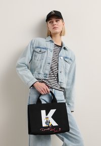 Sac fourre-tout noir avec un "K" blanc et un graphique d'un personnage de dessin animé, doté de petites poignées. Le modèle porte une veste en denim et une chemise rayée.