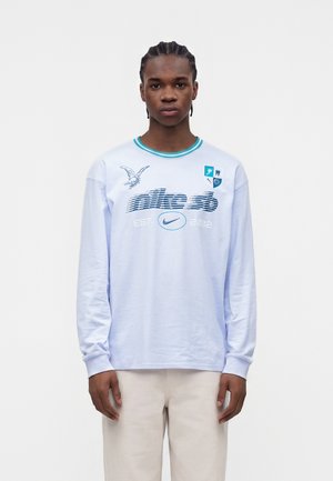 Mladý muž s vrkočmi, oblečený v bielej dlhoruká Nike SB košeli s modrými logami a béžovými nohavicami, stojaci pred jednoduchým pozadím.
