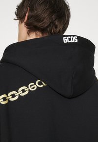 Felpa nera con cappuccio caratterizzata da una grafica di una catena dorata e il testo "GCDS" sulla spalla. Tessuto morbido con una vestibilità rilassata e un ampio cappuccio accogliente.