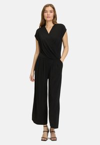 Zwarte jumpsuit met een gewikkelde v-hals, korte mouwen, brede broekspijpen en zijzakken; gemaakt van zachte, luchtige stof. Sandalen met dunne bandjes.