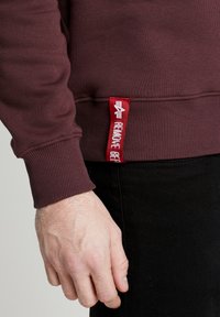 Nærbillede af en persons hånd tæt ved kanten af en vinrød sweatshirt med et rødt mærke, der viser en hvid pil og tekst på siden.
