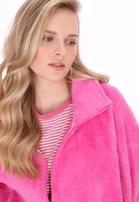 Flauschige rosa Jacke mit Reißverschluss und hohem Kragen, die über einem rosa-weiß gestreiften Hemd getragen wird. Weiche Textur, lässiges Design und lebhafte Farbe.