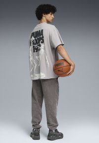 Camiseta gris de gran tamaño con gráfico de esqueleto y texto "PUMA HOOPS". Lleva puesta con pantalones de chándal grises y zapatillas negras, sosteniendo un baloncesto.