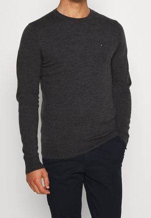 Mann trägt einen dunkelgrauen Langarmpullover mit kleinem Logo auf der Brust, kombiniert mit dunklen Hosen, Hand in der Tasche.