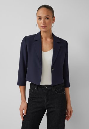 Vrouw die een marineblauw cropped blazer draagt over een wit topje en zwarte jeans, staand tegen een effen lichtgrijze achtergrond.