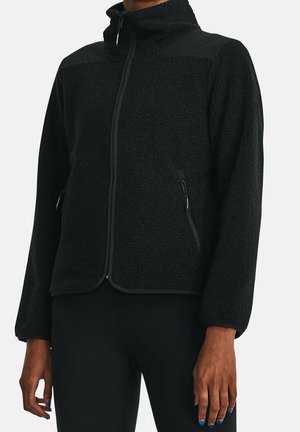 Osoba nosząca czarną polarową kurtkę z pełnym zamkiem błyskawicznym i zasuwanymi kieszeniami bocznymi, w połączeniu z czarnymi spodniami na jasnym tle.