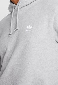 Primer plano de una persona que lleva una sudadera Adidas gris claro con cordones y un pequeño logo Adidas blanco en el pecho.