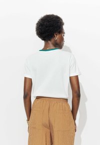 T-shirt blanc à manches courtes avec un détail de col vert, associé à un pantalon marron à rayures fines. Le tissu semble lisse et décontracté.