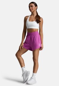 Tætsiddende hvid sports-bh, kombineret med løse lyserøde shorts med elastisk talje og logo. Bærer hvide træningssko og sokker.