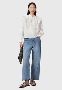 Camicia bianca con colletto arruffato e dettagli in pizzo intricati, abbinata a jeans a gamba larga blu e scarpe ballerine bianche e nere con motivo.