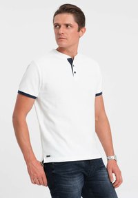 Bílý polo shirt s krátkými rukávy a námořnickým modrým límcem a knoflíky, vyrobený z strukturované látky s bočními detaily. Nosený s tmavými džíny.