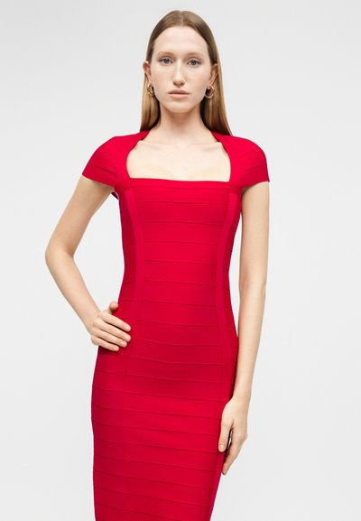 Žena s rovnými blond vlasy, oblečená v těsně přiléhajících červených šatech s krátkými rukávy a hranatým výstřihem, stojící před jednobarevným pozadím.