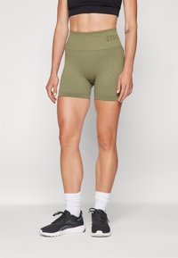 Olivgrüne, gerippte Active Shorts mit hohem Bund, auf der Vorderseite mit dem Wort "STRONG" bedruckt. Getragen mit schwarzen Sportschuhen und weißen Socken.