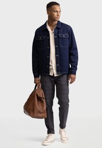 Giacca in jeans blu navy con bottoni, camicia color crema chiaro, pantaloni a scacchi in toni scuri, sneaker beige, holding a brown leather bag.