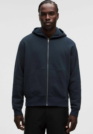 Mann mit geflochtenem Haar, der einen dunkelblauen Kapuzenpullover mit Reißverschluss und schwarze Hosen trägt, steht vor einem schlichten hellen Hintergrund.