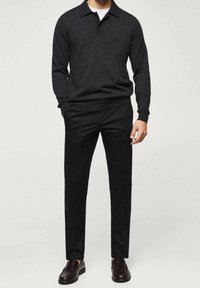 Mann trägt einen dunkelfarbenen Langarm-Polopullover, schwarze Slim-Fit-Hosen und dunkelbraune Loafers, steht vor einem schlichten Hintergrund.