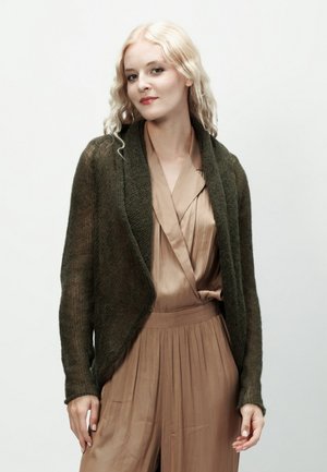 Młoda kobieta o długich blond włosach, ubrana w ciemnozielony dziergany kardigan na beżowej jedwabnej bluzce i plisowanych spodniach, delikatnie się uśmiecha.
