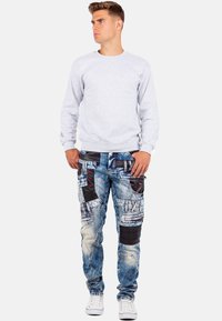 Sweatshirt gris à col rond, associé à un jean en denim bleu déchiré avec des accents noirs, plusieurs poches et des détails de couture. Baskets blanches.