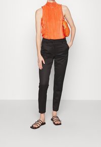 Pomarańczowy plisowany top bez rękawów z wysokim kołnierzem, zestawiony z czarnymi, dopasowanymi spodniami i płaskimi sandałami na paski. Dopełniony teksturowaną pomarańczową torbą.