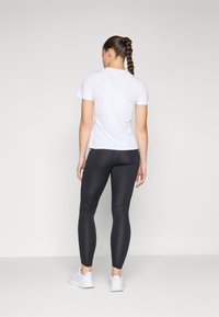 Weißes T-Shirt mit kurzen Ärmeln, kombiniert mit schwarzen Leggings. Die Leggings haben ein figurbetontes Design und eine glatte Textur. Die Fußbekleidung sind hellfarbige Sportschuhe.