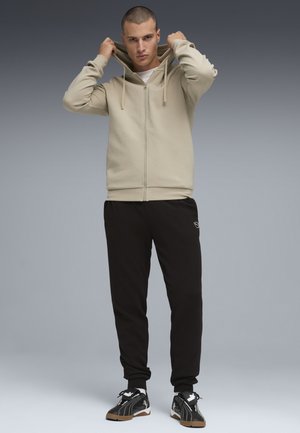 Junger Mann, der eine beige Sweatshirtjacke mit Reißverschluss, schwarze Jogginghosen und schwarze Turnschuhe trägt, hält den Kragen der Kapuzenjacke mit beiden Händen vor grauem Hintergrund fest.