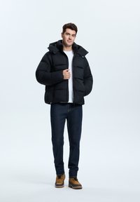 Schwarze Pufferjacke mit Kapuze, ausgestattet mit einem gesteppten Design und einem Reißverschluss, getragen über einem weißen Hemd und kombiniert mit dunklen Jeans und braunen Stiefeln.